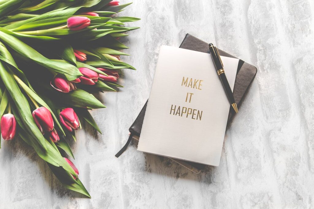 Loo oma unistuste sündmus A notebook with 'Make It Happen' inscription beside a bouquet of pink tulips, capturing inspiration.