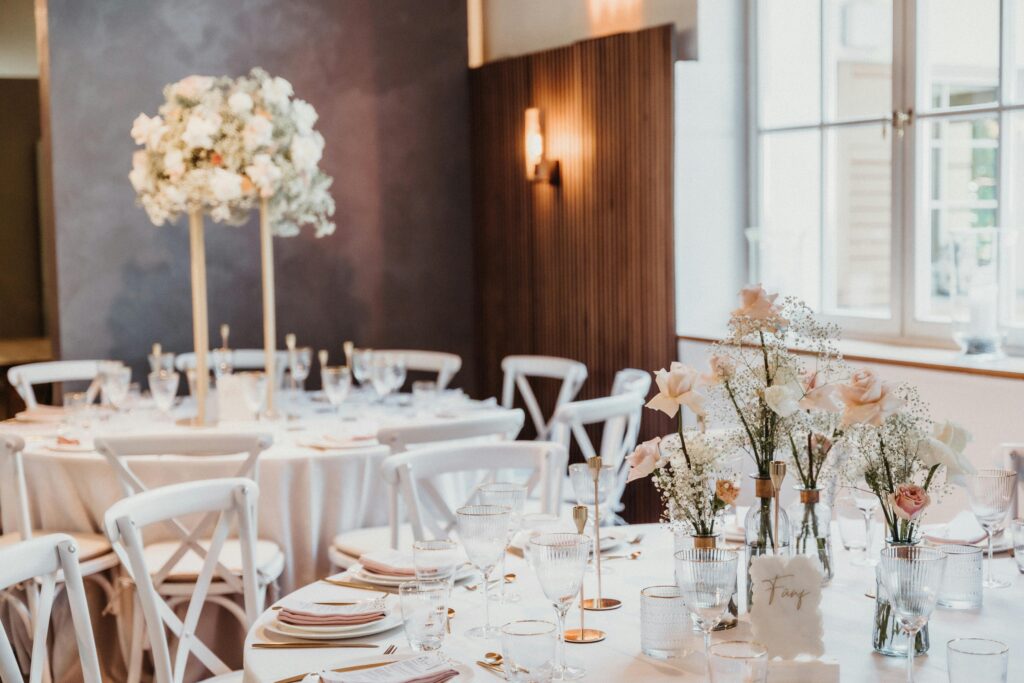 pexels-photo-19351562-19351562 Luxurious wedding setup featuring white tablecloths, floral centerpieces, and elegant tableware in a bright venue.